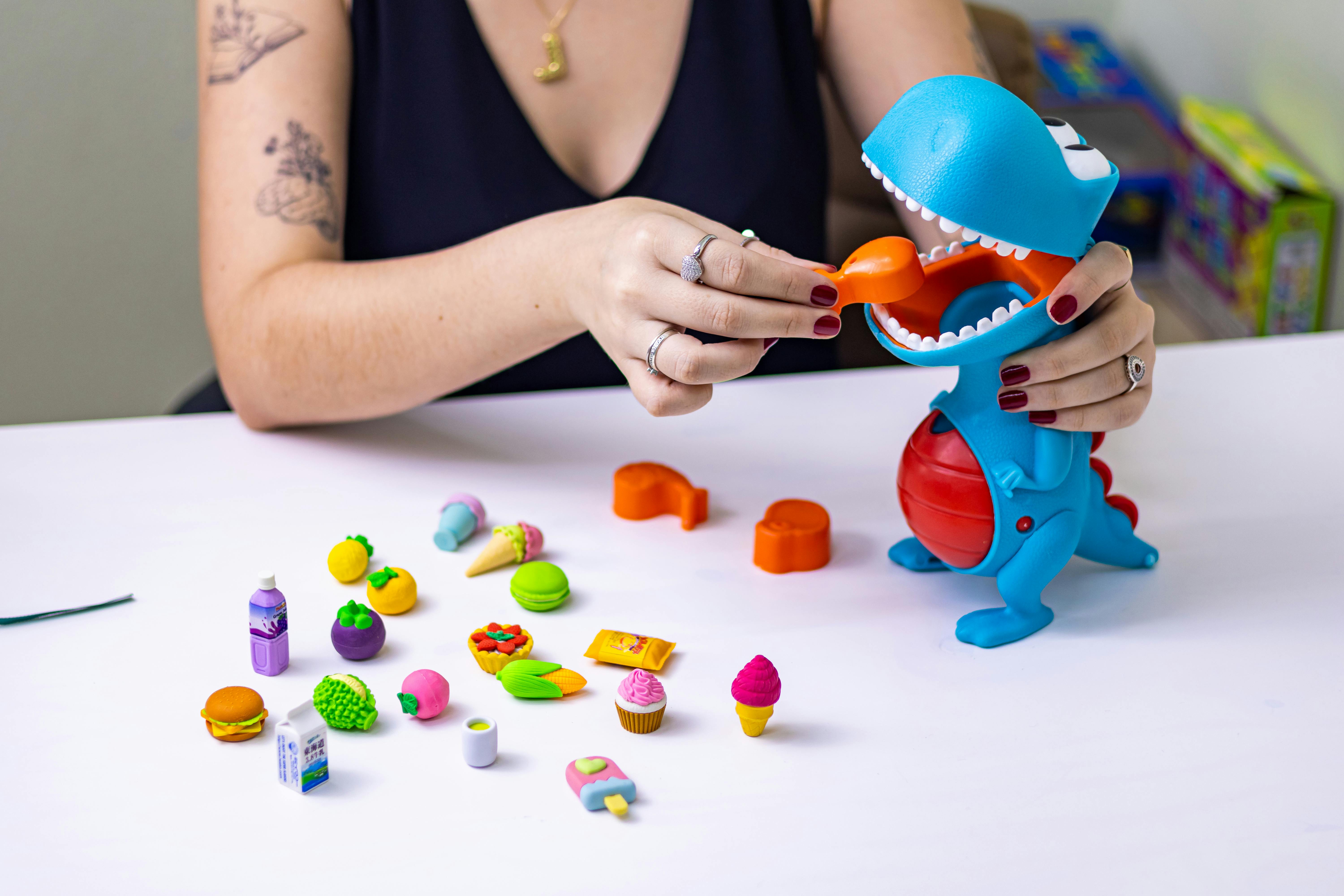 pexels-photo-29803755-29803755 Woman interacting with a toy dinosaur and colorful miniature foods on a table.