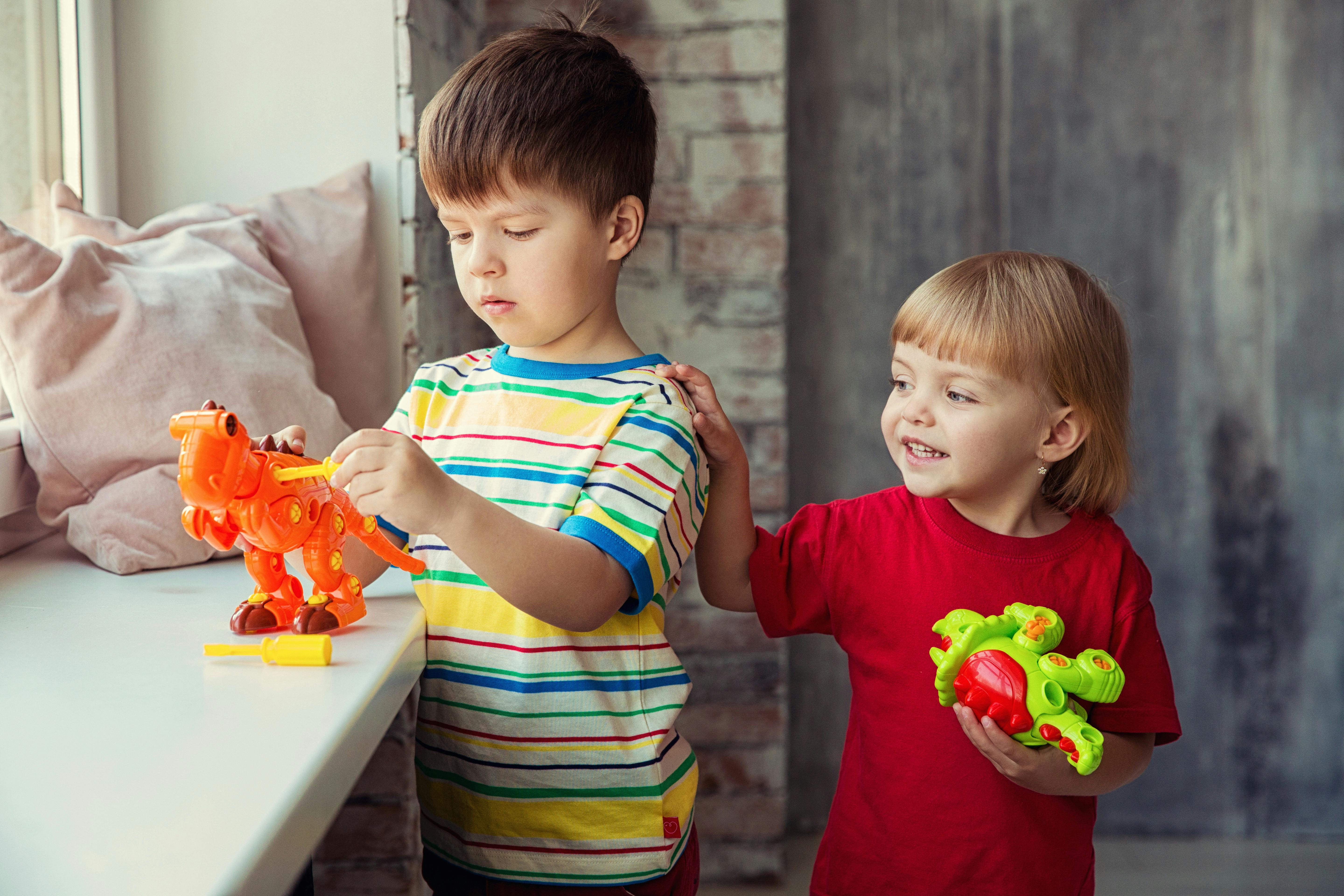 pexels-photo-6139568-6139568 Two kids playing happily with colorful dinosaur toys by a window indoors.